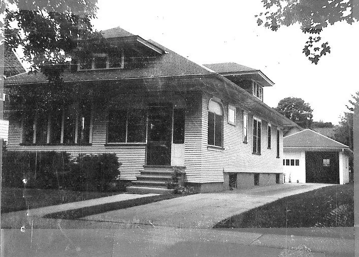 Black-and-white photo of a single-story bungalow with a covered front porch, driveway, and detached garage.