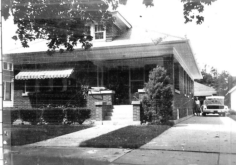 Black-and-white photo of a brick bungalow with a front porch and awning, driveway with a parked car and two people.