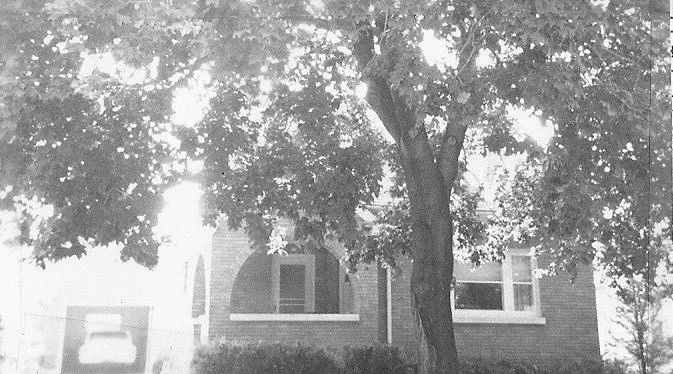 Black-and-white photo of a large tree partially obscuring the front of a brick house with an arched porch and windows.