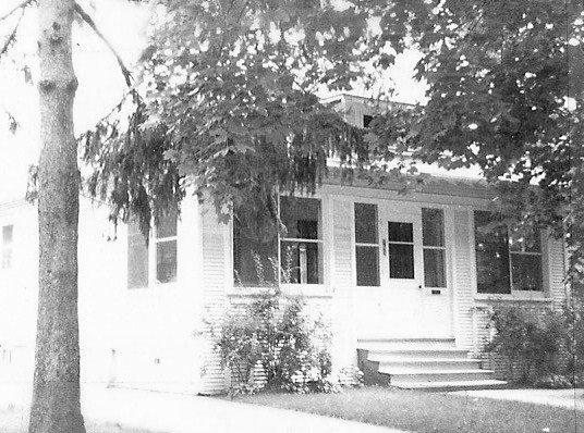 Black-and-white photo of a small bungalow-style house with an enclosed front porch, steps up to the door, and large trees partially shading the yard.