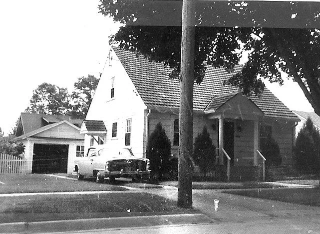 Black-and-white photo of a small gabled house with a detached garage, a vintage car in the driveway, and a utility pole and trees in the front yard.