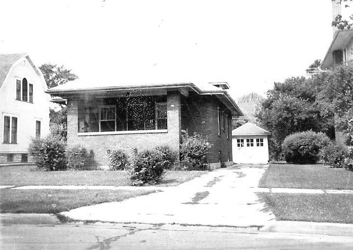 Black-and-white photograph of a single-story brick bungalow with a covered front porch, driveway leading to a detached garage, and shrubs on a residential lot.