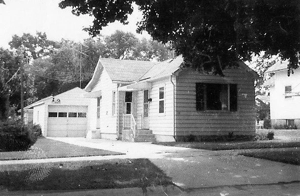 Black-and-white photo of a small two-story bungalow with window awnings, shrubs along the front, and a detached garage visible at left.