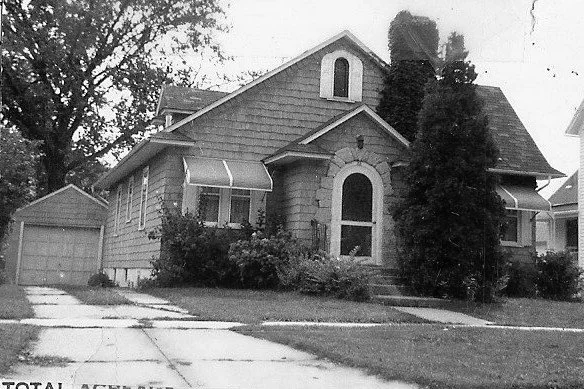 Black-and-white photo of a single-family bungalow with shingled siding, arched front door, awnings over windows, tall shrubs, and a detached garage.