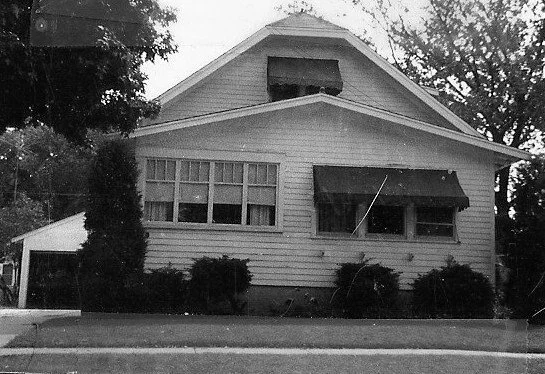 Black-and-white photo of a single-story bungalow with awnings over the front windows, shrubs along the foundation, and a garage at left.