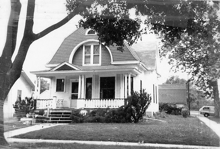 Black-and-white photo of a two-story house with an arched dormer, front porch, and large tree in the foreground.