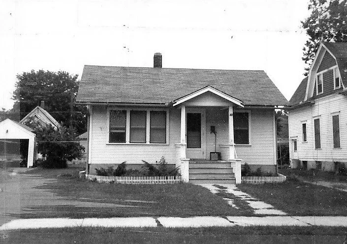 Black-and-white photo of a single-story bungalow with a small covered front porch, three front windows, low picket fence, and driveway beside neighboring houses.