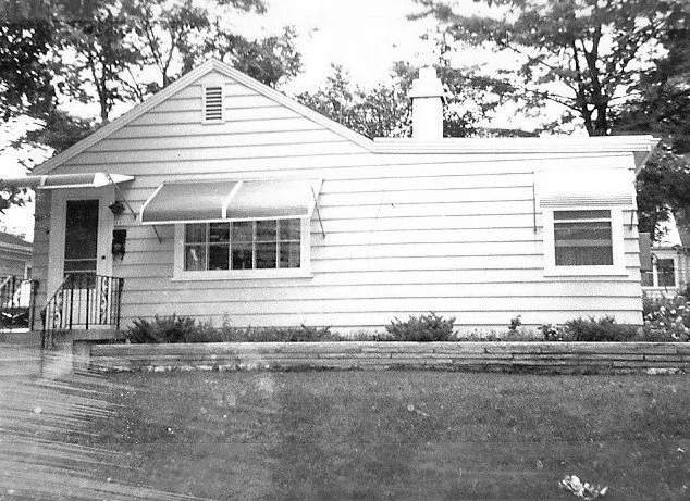 Black-and-white photo of a single-story suburban house with horizontal siding, a front window awning, small porch steps, and a grassy front lawn.