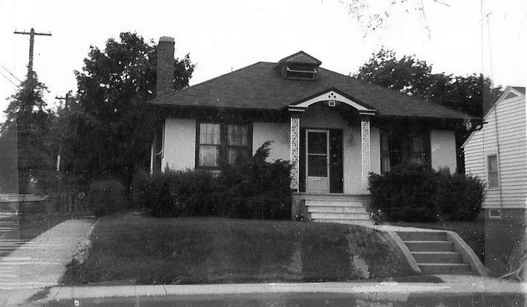 Black-and-white photo of a single‑story bungalow with a small front porch, steps up a sloped lawn, and shrubs along the facade.