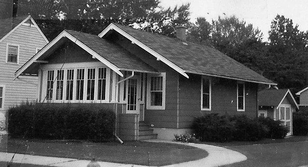 Black-and-white photo of a single-story Craftsman-style bungalow with an enclosed front porch, shrubs, curved sidewalk, and neighboring houses.