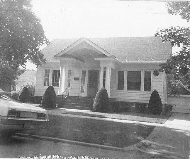 Black-and-white photo of a one-story bungalow with a covered front porch supported by columns, trimmed shrubs at the entrance, and a car parked at the left foreground.