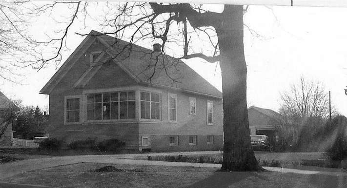 Black-and-white photo of a one-story bungalow with an enclosed front porch, a large tree in the front yard, and a driveway leading to a garage with a parked car.