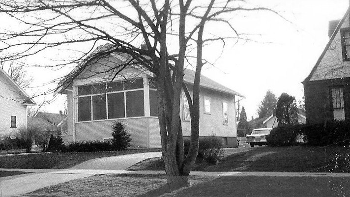 Black-and-white photo of a single-story house with an enclosed front porch, a leafless tree in the foreground, driveway, and a parked car beside the house.