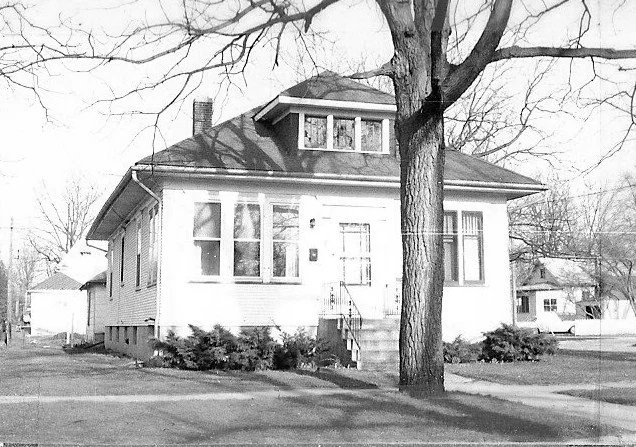 Black-and-white photo of a single-story bungalow with a dormer, front steps, shrubs, and a large leafless tree in the front yard.