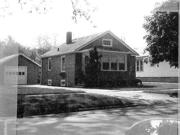 Black-and-white photo of a single-story brick bungalow with a front enclosed porch, detached garage to the left, and a tree-lined yard.