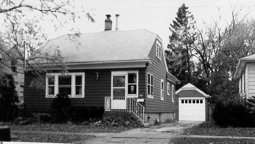 Black-and-white photo of a small single-story suburban house with a front porch, steps, driveway and a detached one-car garage, surrounded by trees.