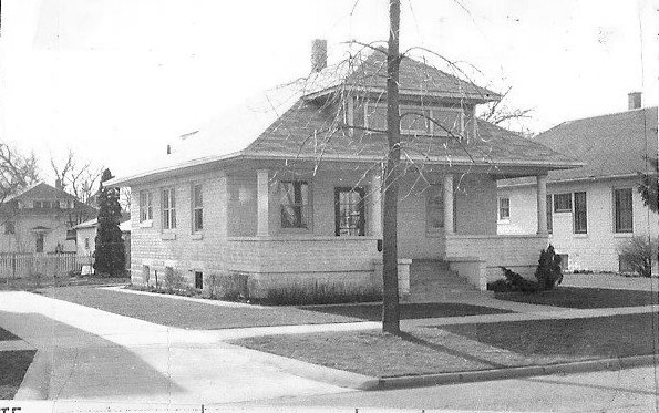 Black-and-white photo of a single-story Craftsman-style bungalow on a residential corner lot with a small front porch, a bare tree and a utility pole in the foreground.