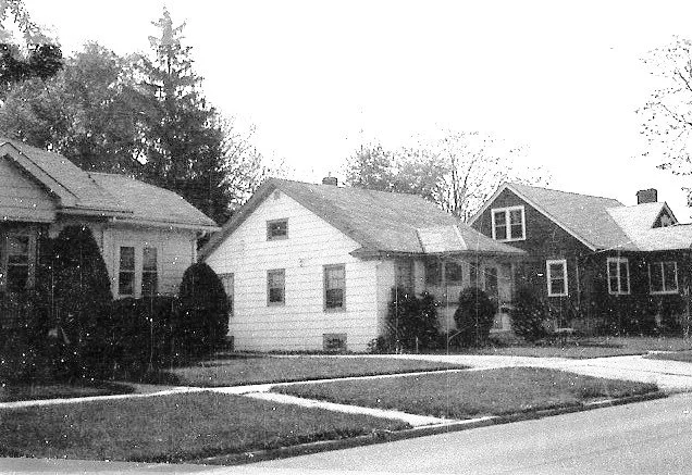 Black-and-white photo of a quiet suburban street with three small houses, front lawns, and a sidewalk.