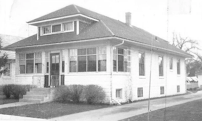 Black-and-white photo of a single-story house with an enclosed front porch, low-pitched hip roof with a small dormer, concrete steps and shrubs in front.