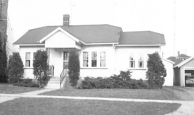 Black-and-white photo of a single-story bungalow with a central covered porch, flanked by windows, shrubs and two tall evergreens, and a driveway to a detached garage on the right.