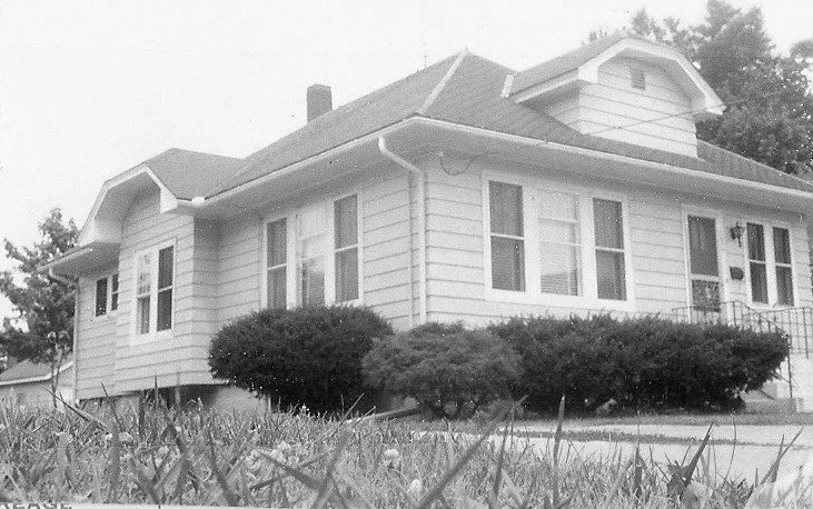 Black-and-white photo of a single-story bungalow with front shrubs and grass in foreground.