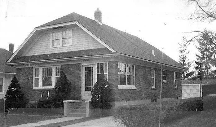 Black-and-white photo of a one-and-a-half-story brick bungalow with front steps, small shrubs, and a detached garage.