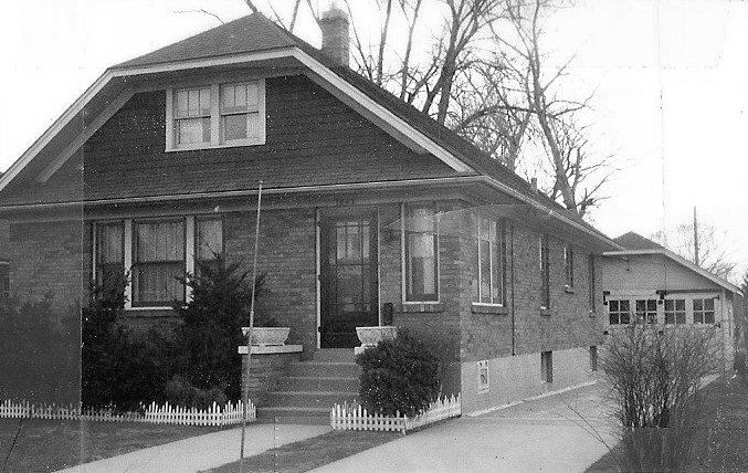 Black-and-white photo of a one-and-a-half-story brick bungalow with front steps, small white picket border, driveway leading to a detached garage, and leafless trees in the background.