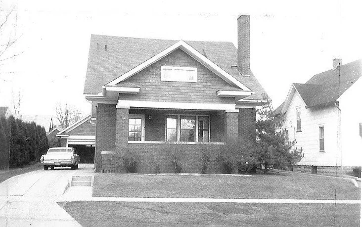 Black-and-white photo of a brick bungalow with covered front porch, chimney, driveway with a parked vintage car.