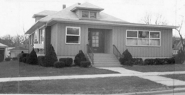 Black-and-white photo of a one-story bungalow with a central front door reached by steps, low shrubs along the foundation, and a sidewalk in front.