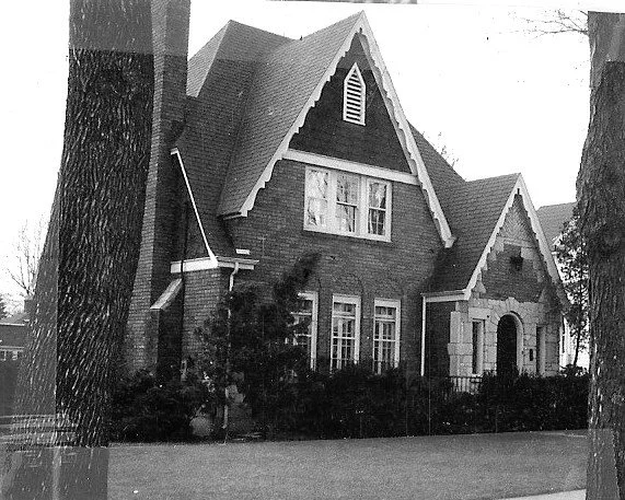 Black-and-white photo of a two-story brick Tudor-style house with steep gables, arched stone entryway, shrubs, and large trees flanking the lawn.