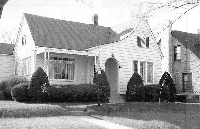 Black-and-white photo of a single-story suburban house with an arched front entry, gabled roof, large front windows and trimmed shrubs.
