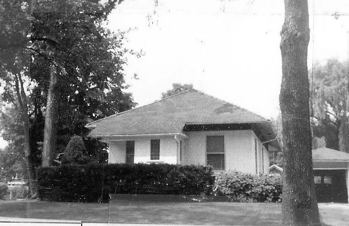 Black-and-white photo of a single-story bungalow with a low hip roof, front hedge, and large trees.