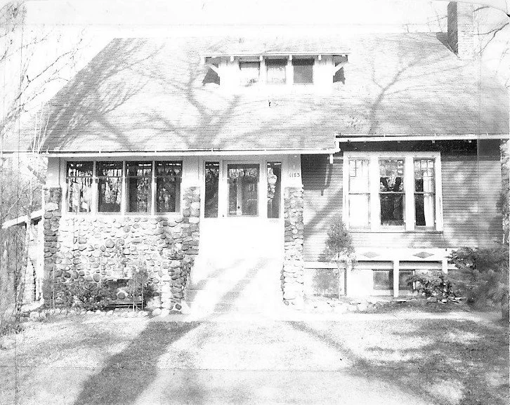 Front view of a vintage bungalow with a stone-pillared enclosed front porch, central steps and door, wide windows, a small dormer, and tree shadows cast across the façade.