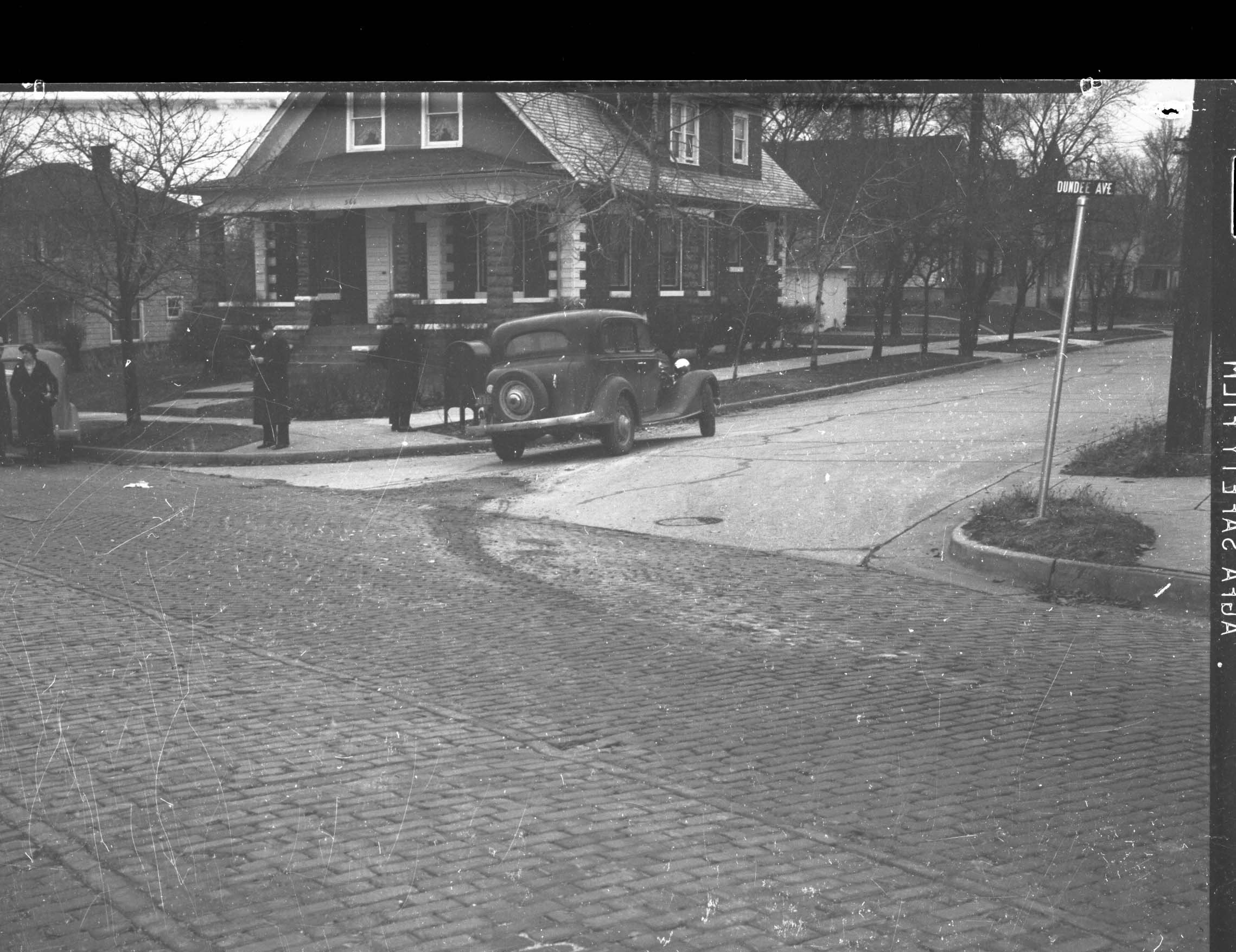 Black-and-white photo of a vintage car parked at a residential corner in front of a two-story house, people on the sidewalk and a "Dundee Ave" street sign.