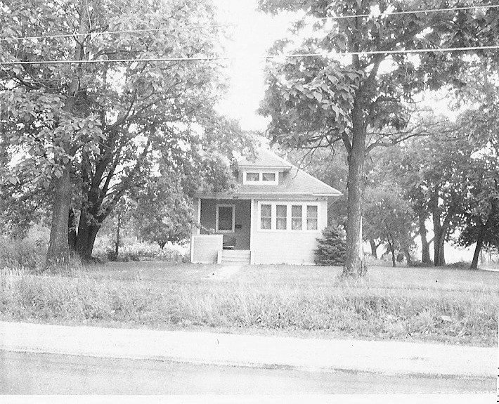 Black-and-white photo of a small bungalow with a front porch, surrounded by large trees and a grassy yard, with a road in the foreground.