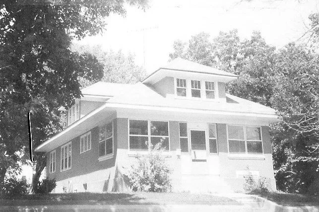 Black-and-white photo of a two-story house with an enclosed front porch, dormer windows, and trees on either side.