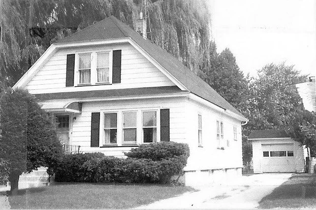 Black-and-white photo of a two-story white house with dark shutters, front shrubs, a driveway, and a detached garage.