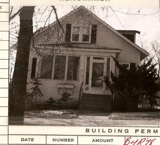 Black-and-white photo of a two-story house with a front enclosed porch, steps up to the door, bare trees and shrubs, and a partial "BUILDING PERM" form visible at the bottom.