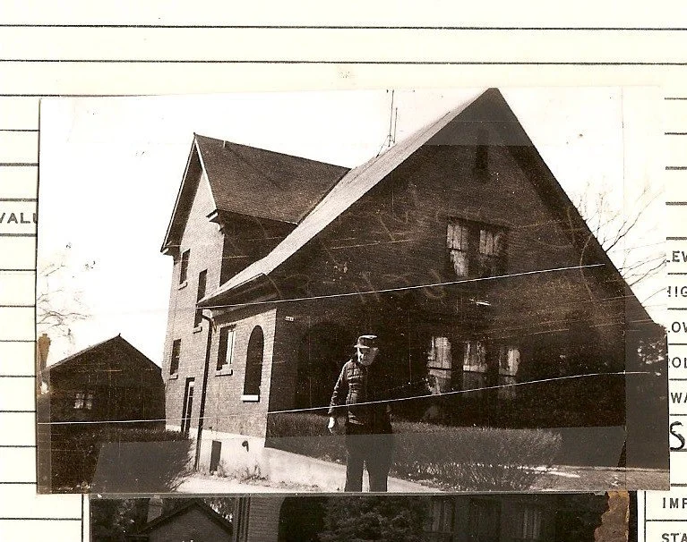 Vintage black-and-white photo of a two-story brick house with a steep gabled roof and covered front porch; a man in a coat and cap stands on the walkway, the print appears pasted onto lined paper.