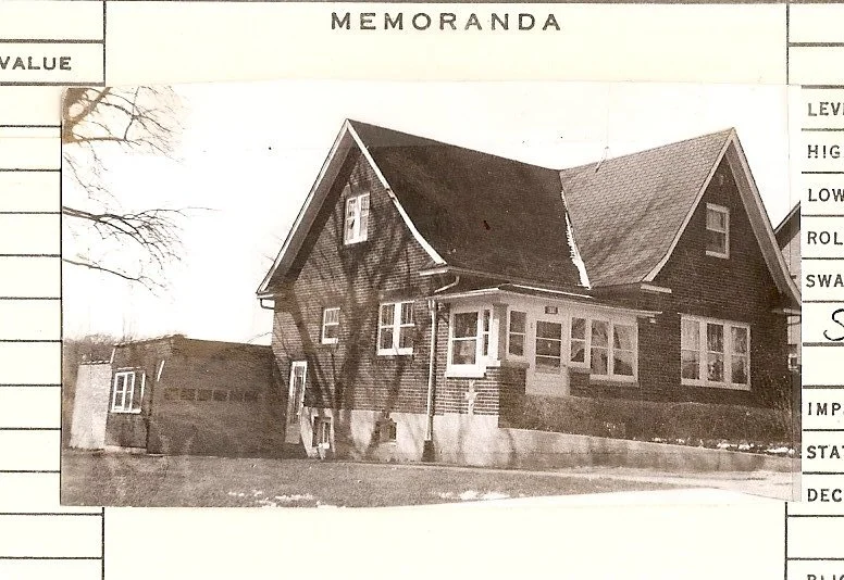 Black-and-white photo of a brick two-story bungalow with steep gabled roof, enclosed front porch and detached garage, mounted on a "MEMORANDA" form.