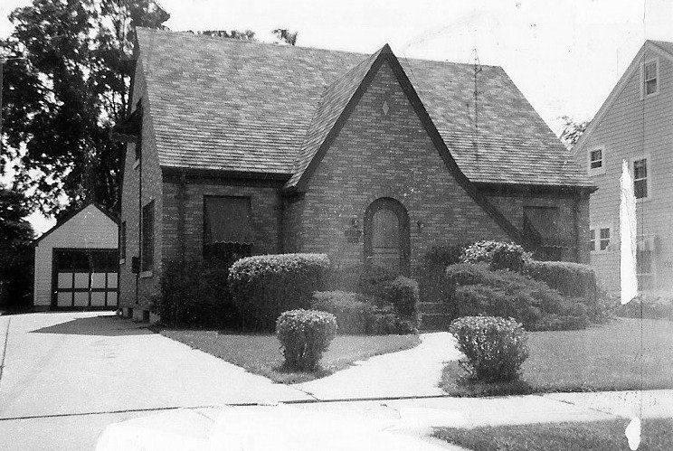 Black-and-white photo of a small brick Tudor-style house with a steep gabled roof, arched front door, trimmed shrubs in the front yard and a detached garage to the left.
