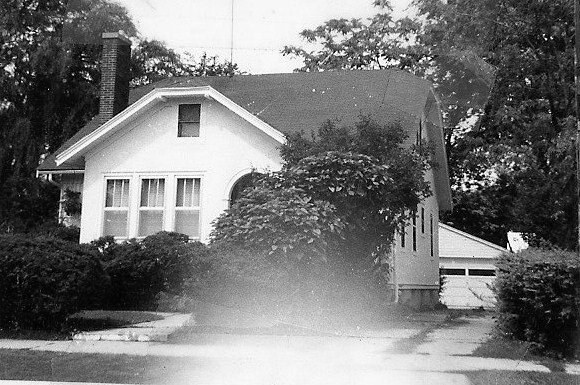 Black-and-white photo of a small bungalow-style house with a front porch window, overgrown shrubs, driveway, and a detached garage.