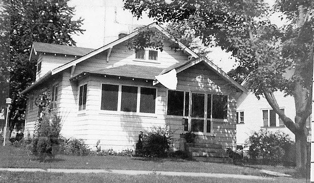 Black-and-white photo of a single-story bungalow with a screened front porch, small flag hanging from the roof, and trees framing the yard.