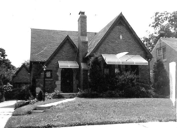 Black-and-white photo of a small gabled brick bungalow with a central chimney, window awnings, shrubs in front, and a sloped lawn.