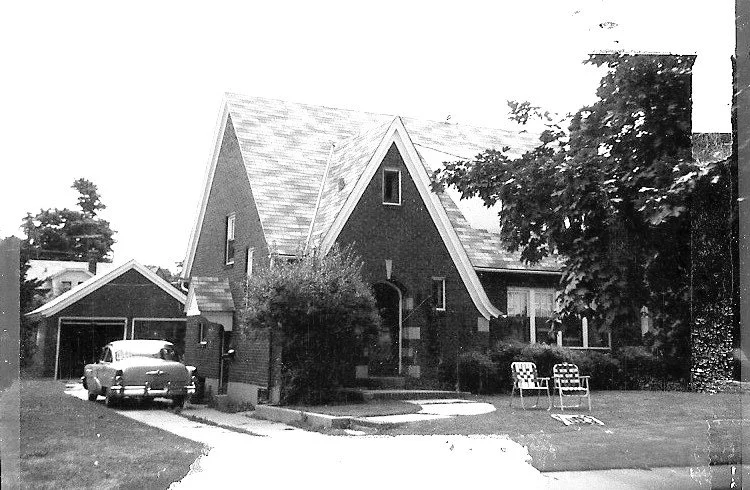 Black-and-white photo of a brick Tudor-style house with steep gables, a vintage car by a detached garage, and two lawn chairs on the front lawn.
