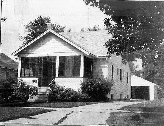 Black-and-white photo of a small single-story bungalow with a screened front porch, steps, driveway leading to a detached garage, and a large tree at the right.