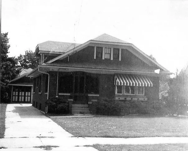 Black-and-white photo of a brick Craftsman-style bungalow with a covered front porch, striped window awning, driveway, and detached garage.