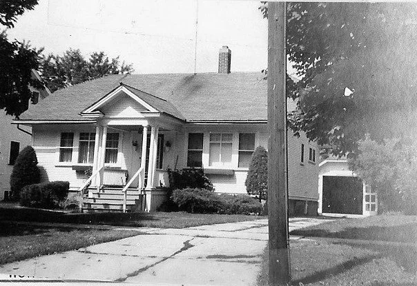 Black-and-white photo of a single-story bungalow with a small front porch, steps and shrubs, a cracked driveway leading to a detached garage, and a utility pole and trees in the foreground.