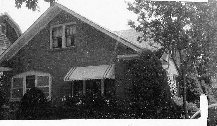 Black-and-white photo of a brick bungalow with a striped awning over the front window, surrounded by trees and shrubs.