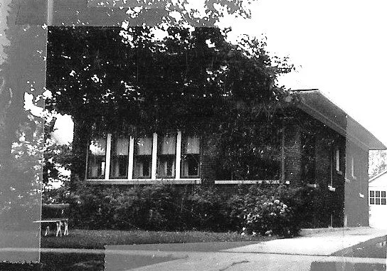 Black-and-white photo of a one-story brick house with five front windows, shrubs and trees around it, and a driveway to the right.